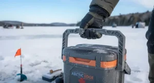 An angler holding the sturdy handle of a portable insulated ice fishing bucket on the snow.